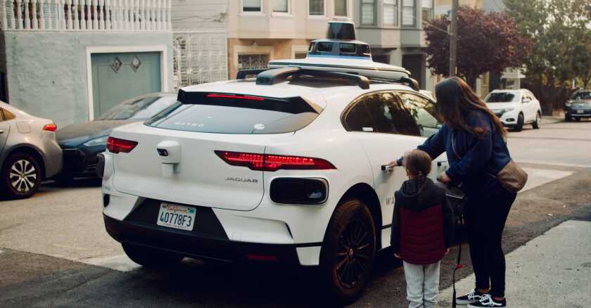 On a city street, a woman and child prepare to enter a Waymo four-door Jaguar vehicle.