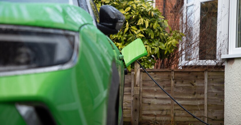 A green-colored electric vehicle faces the camera, charging in front of a wood fence.